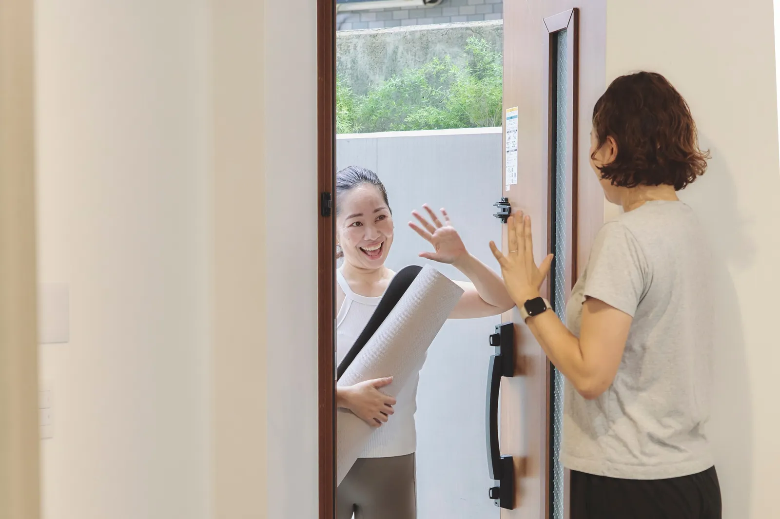 ヨガマットを抱えて玄関から訪れたトレーナーと、手を上げて迎えるお客様